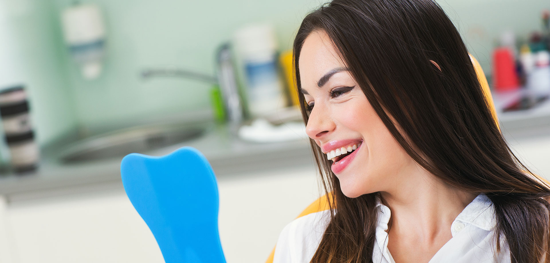 The image shows a woman with a smile, holding an object that resembles a toothbrush or dental tool, in front of her face. She is seated at a countertop with various items behind her, including what appears to be a sink and a cup.