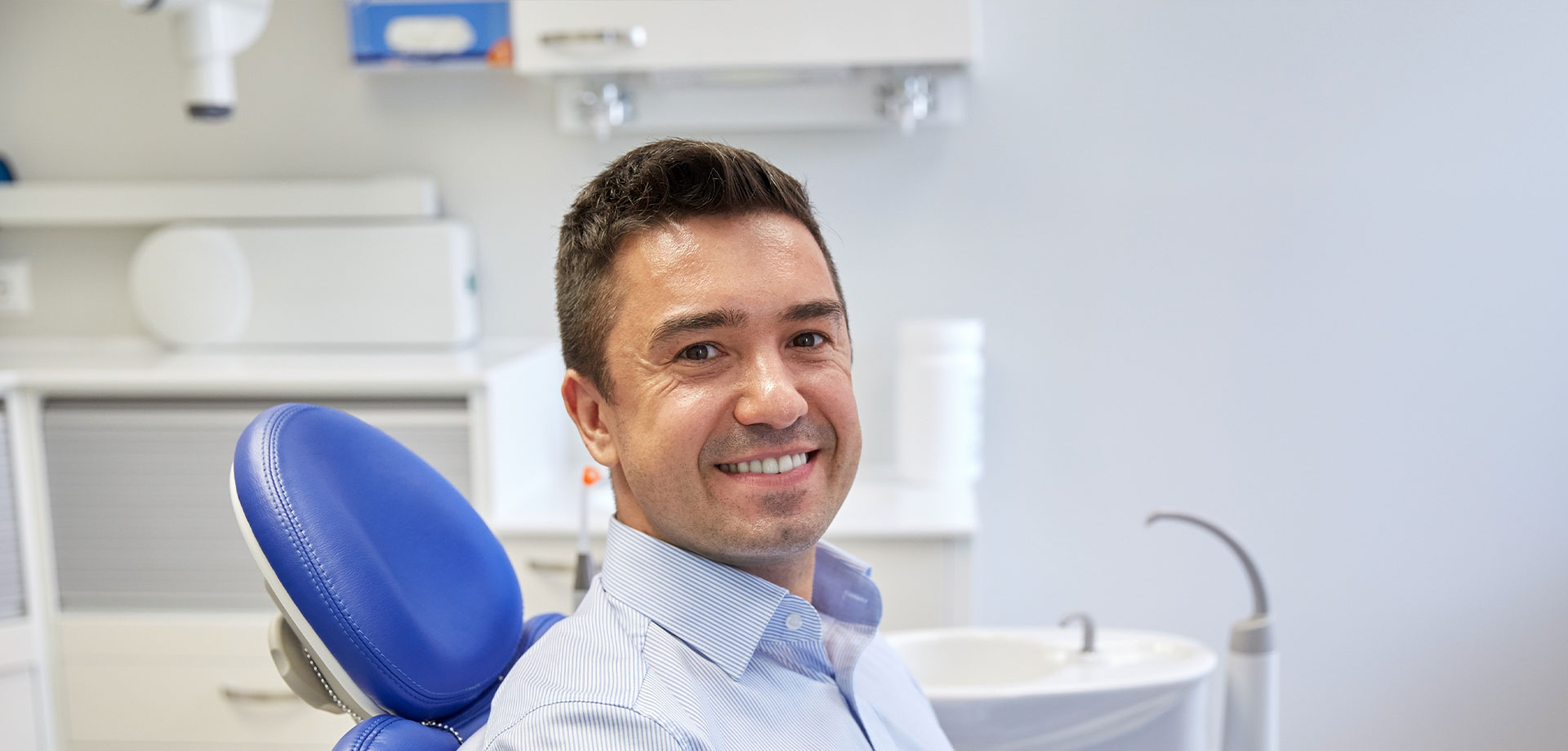 The image shows a man sitting in an office chair, smiling at the camera. He appears to be wearing a white shirt and is situated in front of a desk with a computer monitor on it. Behind him, there s a dental chair, indicating that the setting might be a dental office or a similar professional environment.