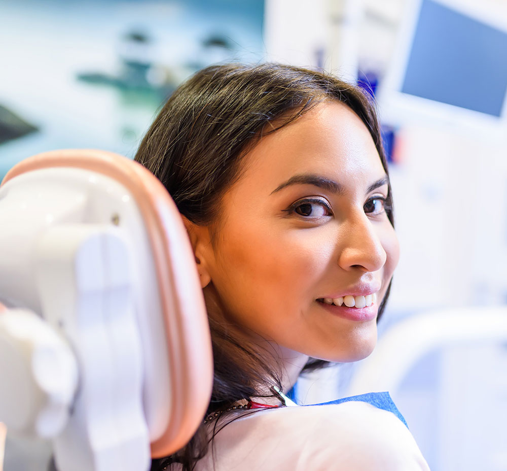 The image shows a woman seated in a dental chair, smiling and looking directly at the camera.