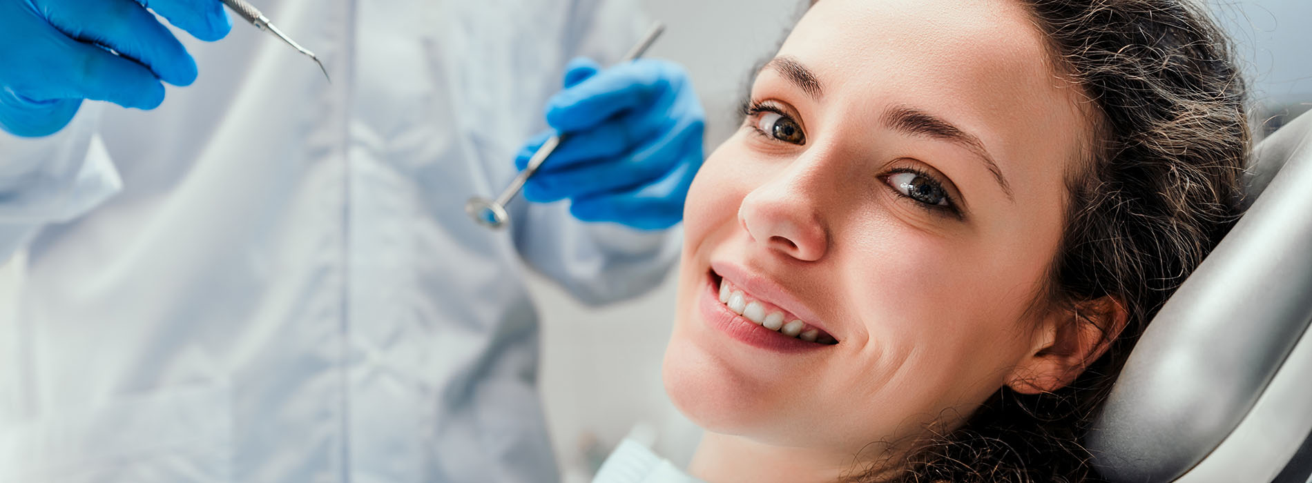 The image shows a woman sitting in a dental chair, receiving dental care from a dentist who is wearing gloves and working with dental instruments.