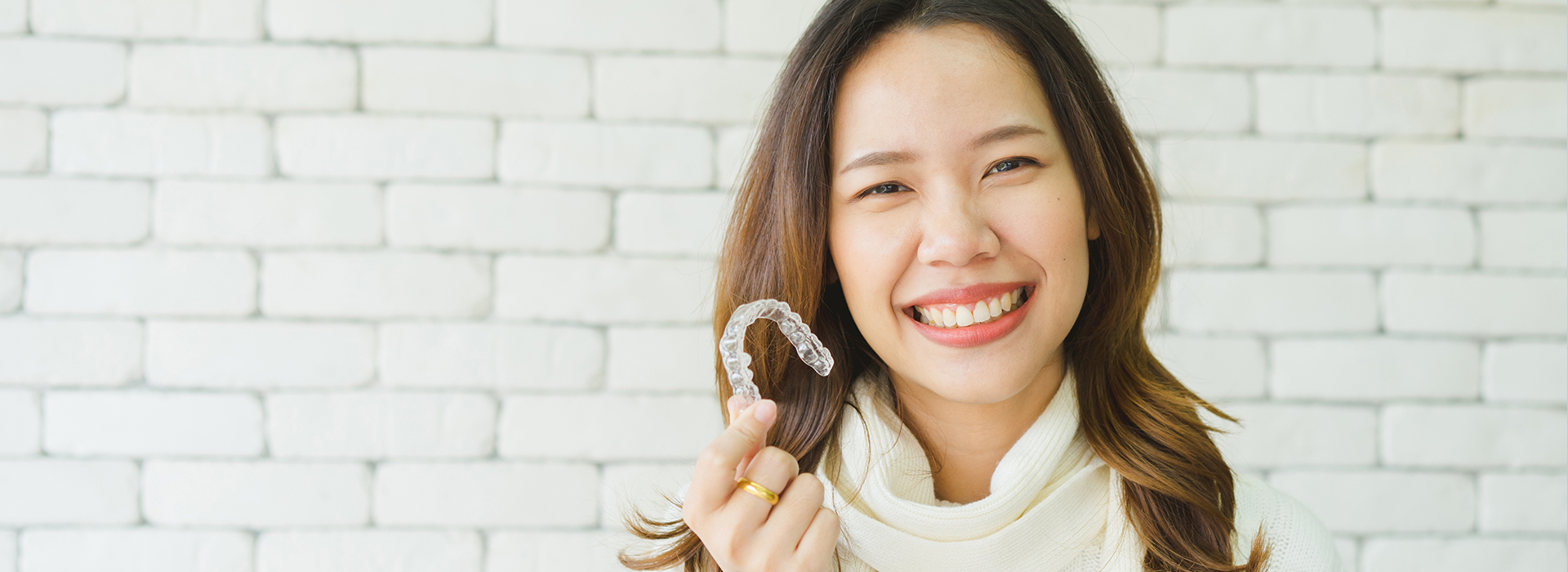 A woman with a smile is holding a ring, set against a brick wall background.